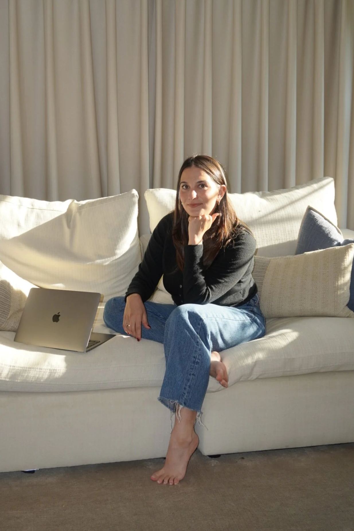 A woman sitting on a white sofa with an open MacBook laptop beside her, in a room with beige curtains, wearing a black sweater and blue jeans, with one leg crossed over the other and her chin resting on her hand.