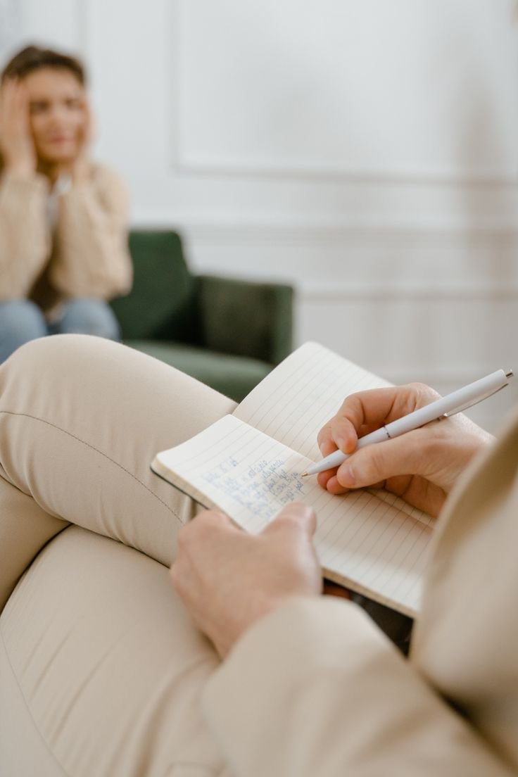 Person sitting on a beige couch taking notes in a small notebook with a pen, with a woman sitting on a green couch in the background, blurred.