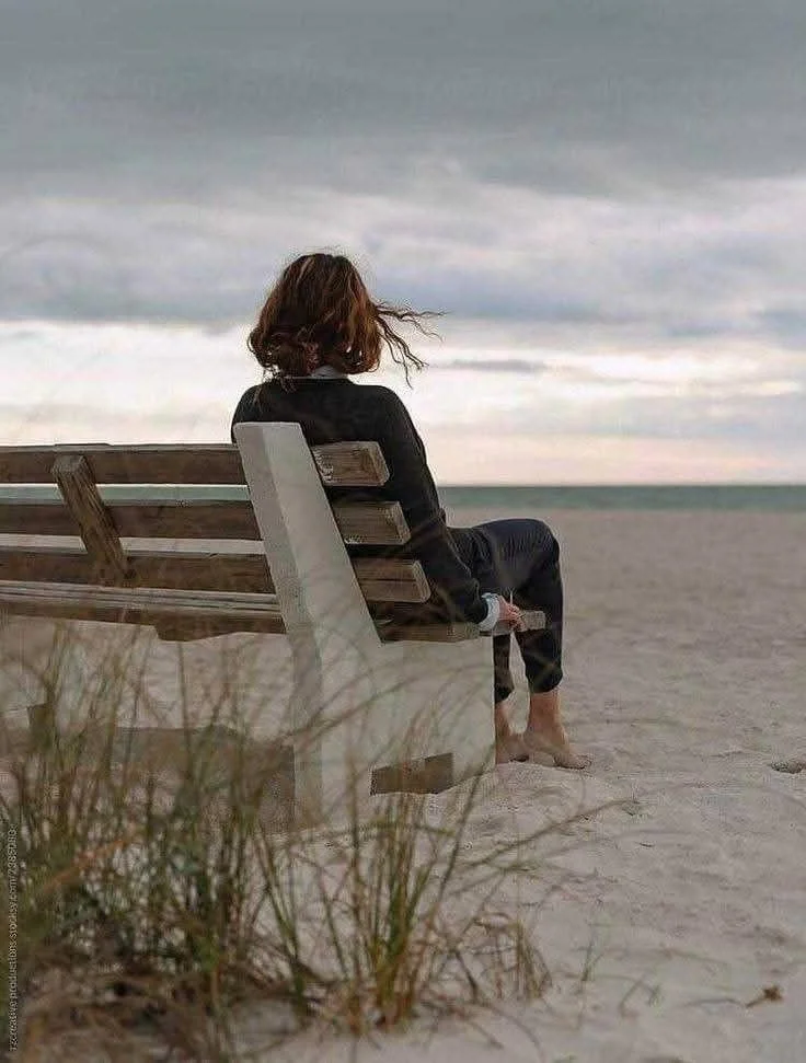A person sitting on a bench on the sandy beach, facing the ocean and looking at the cloudy sky.