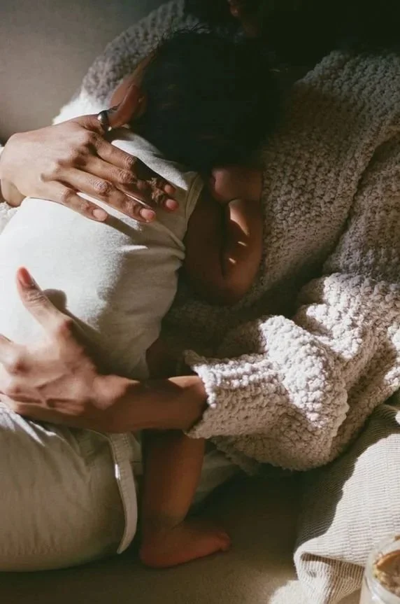 A person holding and cuddling a sleeping child, both lying on a soft surface. The person is wearing a grey, knit sweater, and the child is in a white shirt.