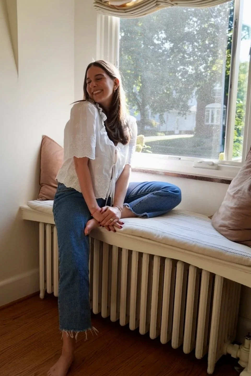 A young woman sitting on a window seat, smiling with eyes closed, wearing a white blouse and blue jeans, during daytime with sunlight streaming through the window.