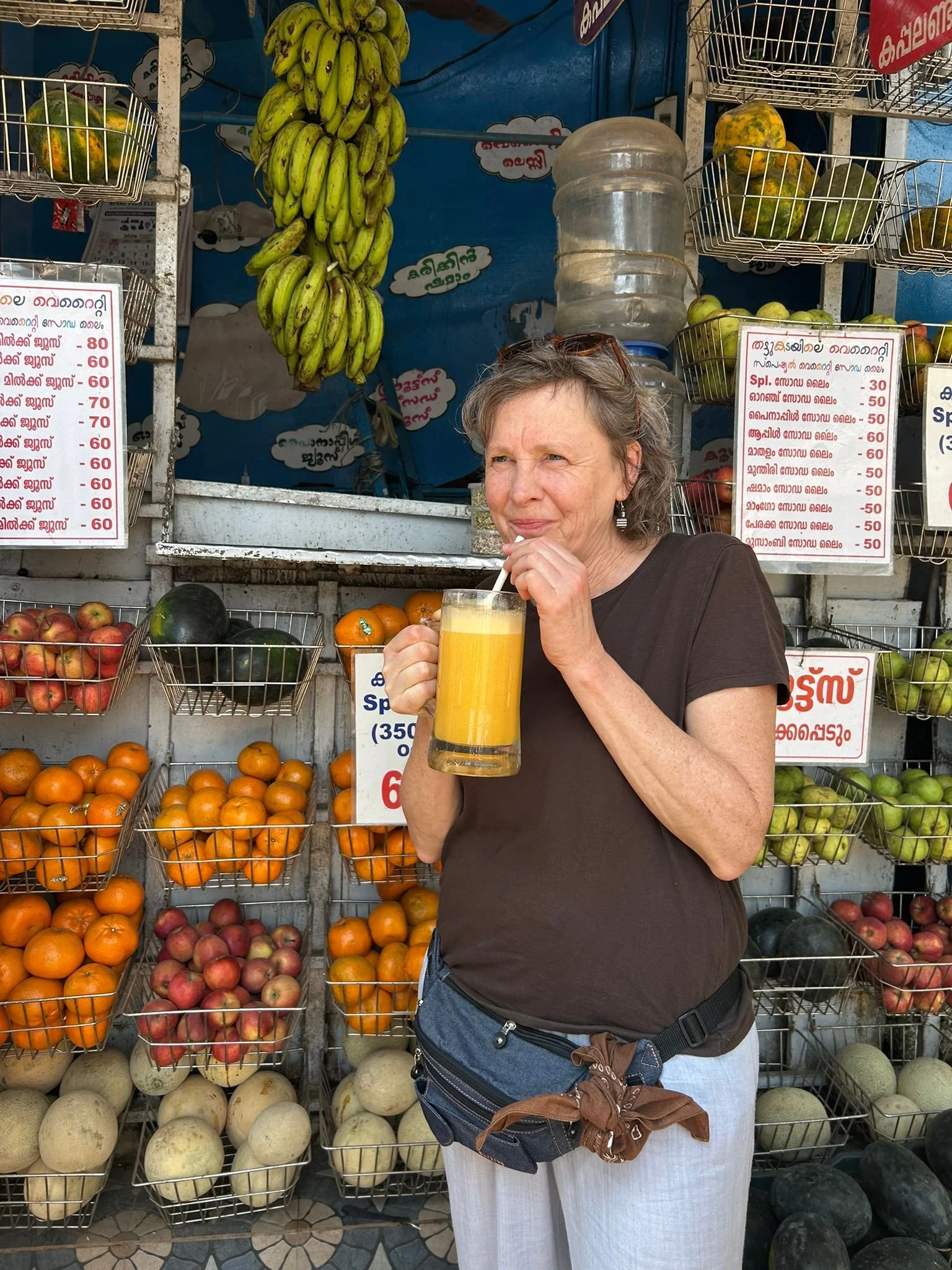 A middle-aged woman with curly gray hair drinking orange juice through a straw at an outdoor fruit stall with various fruits, including bananas hanging overhead, oranges, apples, and melons.