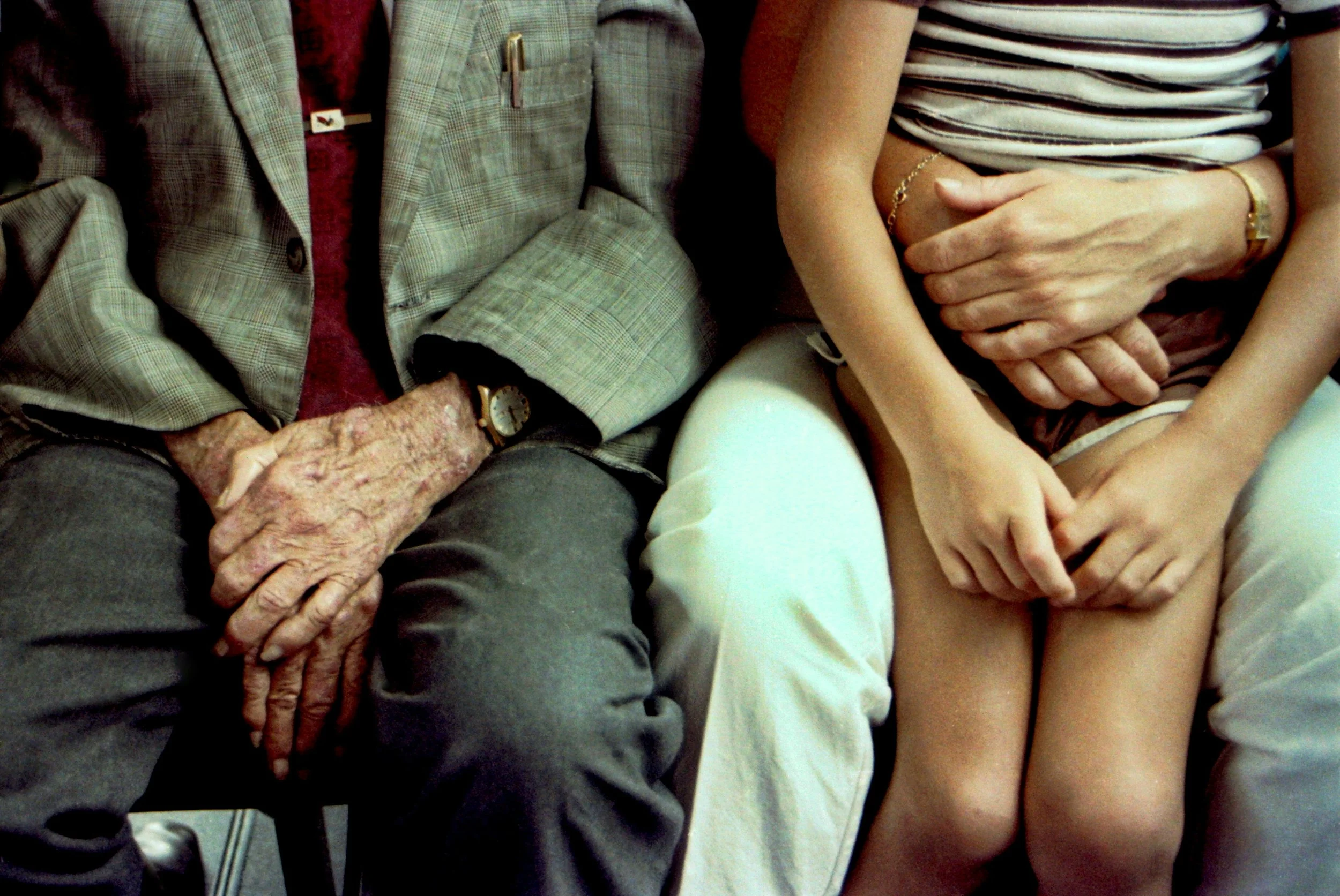 Close-up of two people sitting side by side, showing their hands and legs. One person has aged, wrinkled hands with a watch, and is wearing a gray checked blazer. The other has smoother hands placed over their lap, wearing a striped shirt and beige shorts, with a gold bracelet and watch.