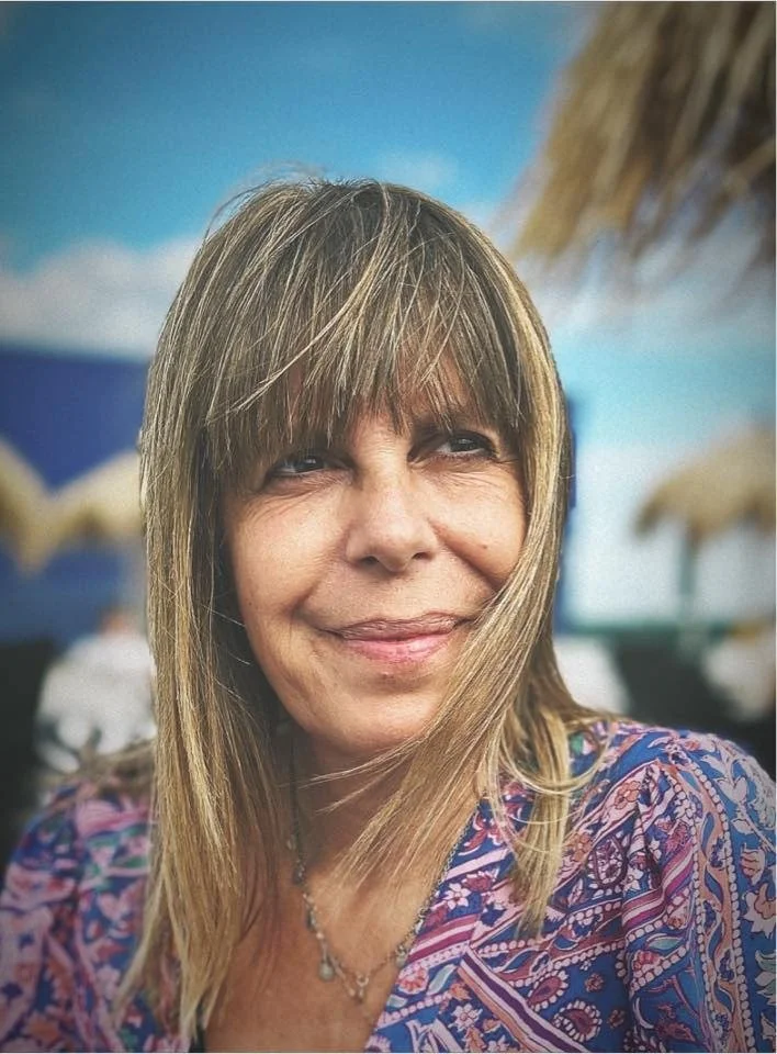 A smiling woman with light brown hair and bangs, wearing a colorful patterned top, outdoors at a beach or tropical location with thatched roof umbrellas in the background.