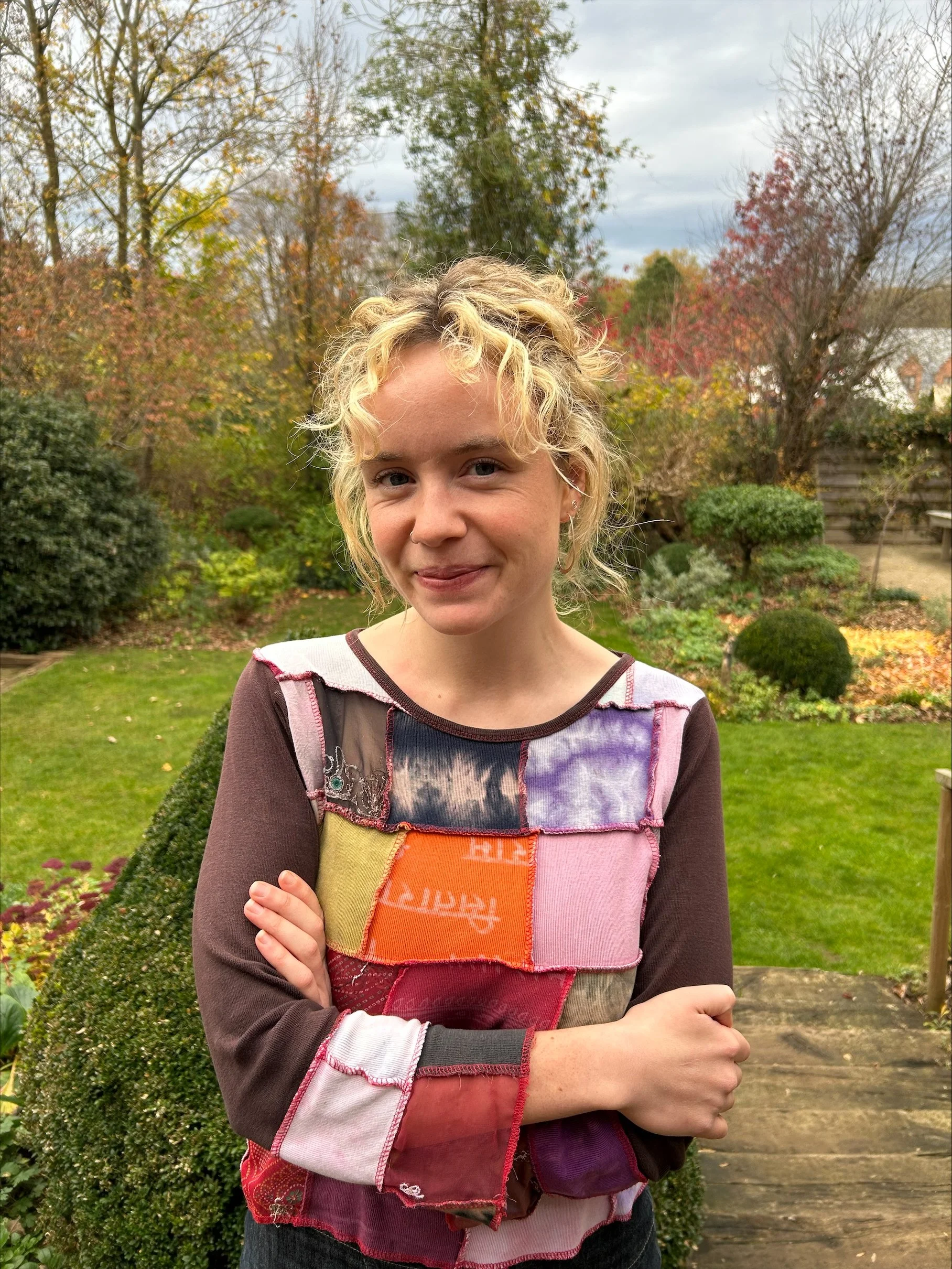 A young woman with curly blonde hair standing in a garden with trees showing fall foliage.