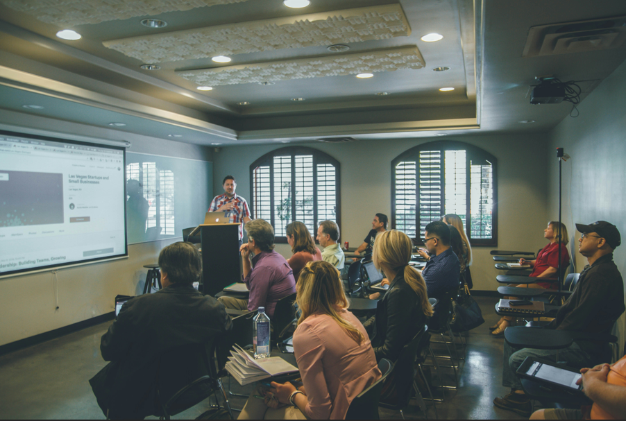 A man giving a presentation to a group of adults in a conference room. The room has large windows with valances, and a projector screen displaying a webpage.