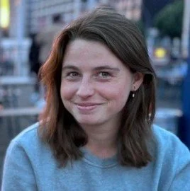 A young woman with shoulder-length brown hair and blue eyes, smiling outdoors in a blue sweatshirt.