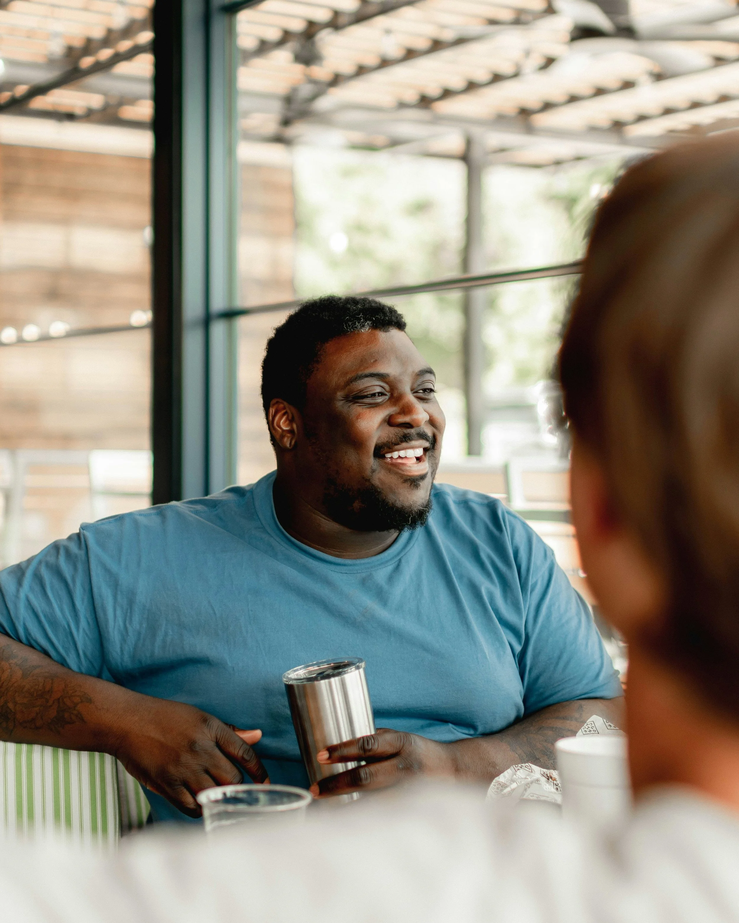 A smiling Black man wearing a blue t-shirt, sitting at a table in a casual setting, holding a stainless steel tumbler, engaged in conversation with a person whose back is partially visible.