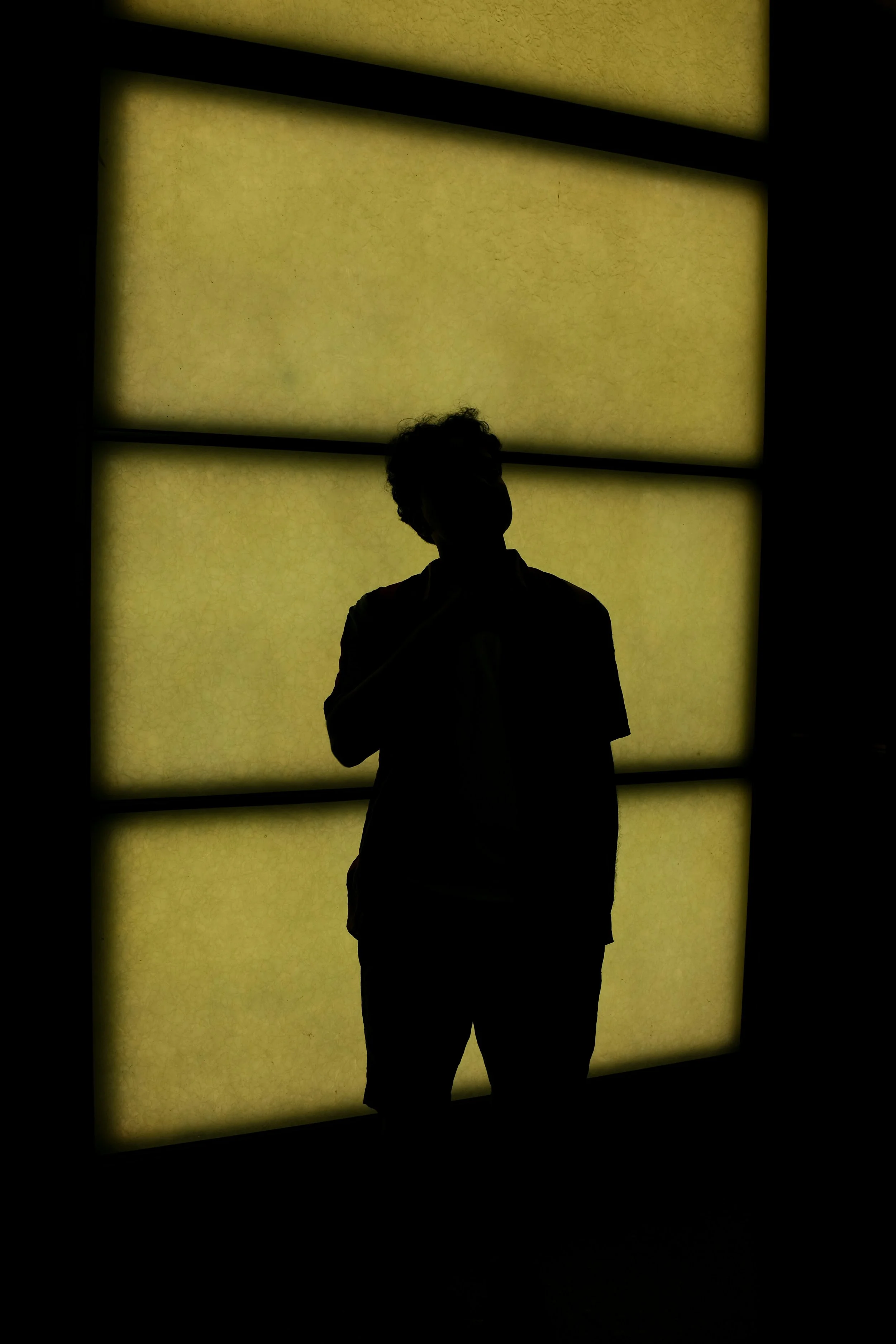 Silhouette of a person standing in front of a backlit yellow window, with their head slightly tilted and one hand near their face.