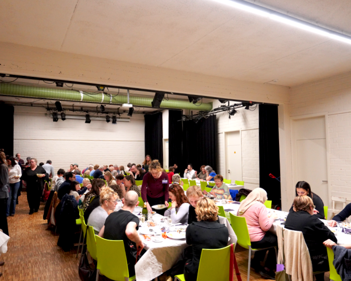 People seated at long tables in a large event hall, engaging in a meal or gathering, with a stage area and black curtains in the background.