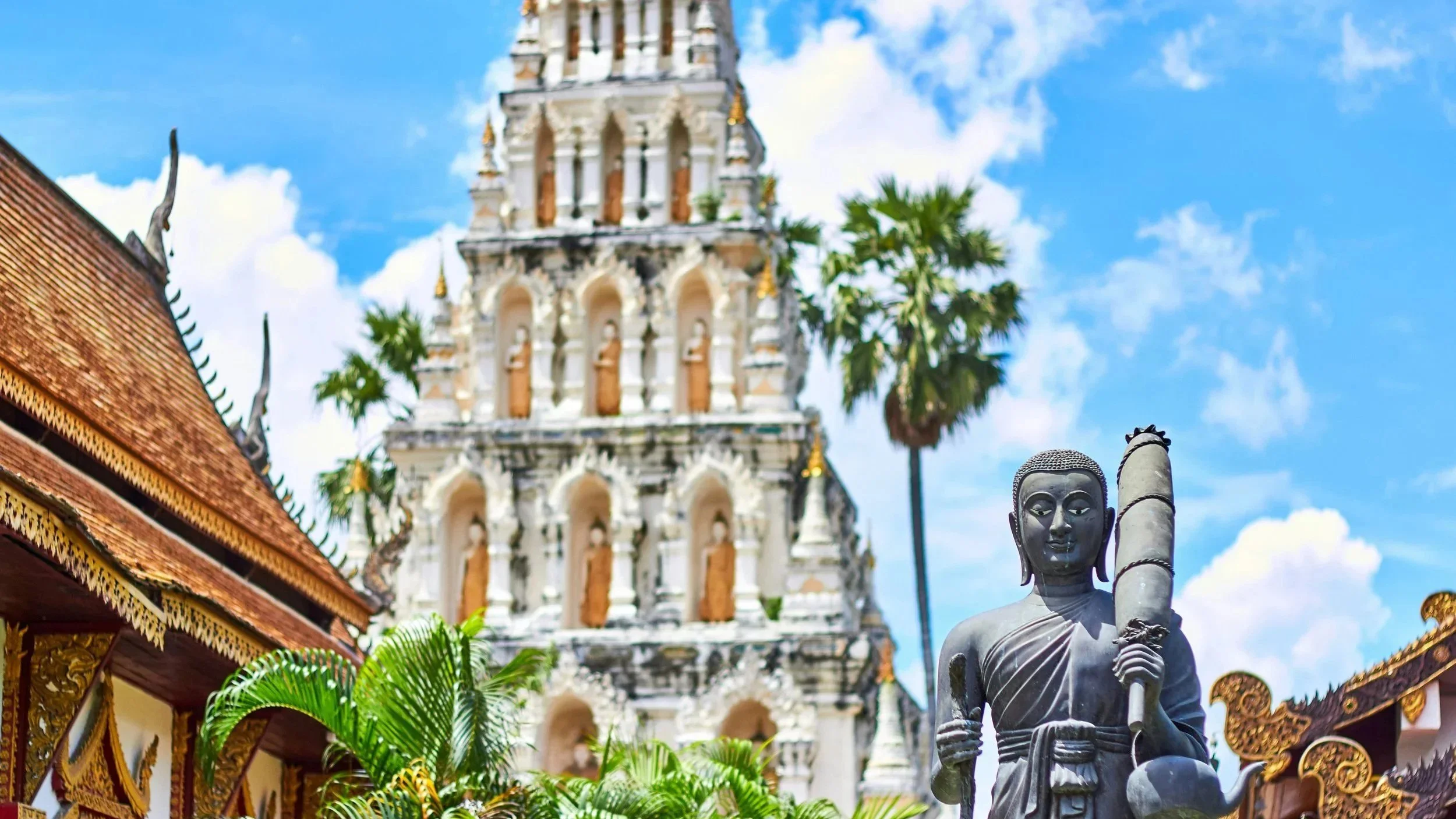 Statue eines Mönchs vor einem traditionellen Tempel mit mehrstöckigem Prinzengarten und Palmen im Hintergrund, Himmel mit Wolken