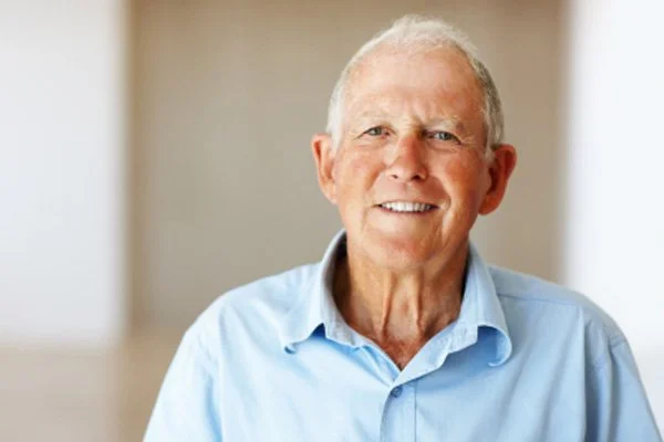 A cheerful older man in a light blue shirt smiles gently while seated indoors.
