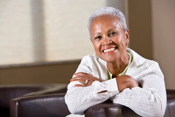 A joyful older woman with short gray hair smiles while relaxing on a sofa.