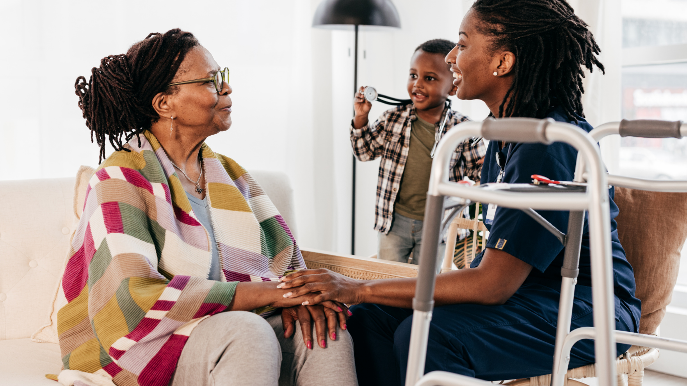 An older woman shares a warm moment with her caregiver as a young child happily plays beside them, creating a caring multigenerational scene.
