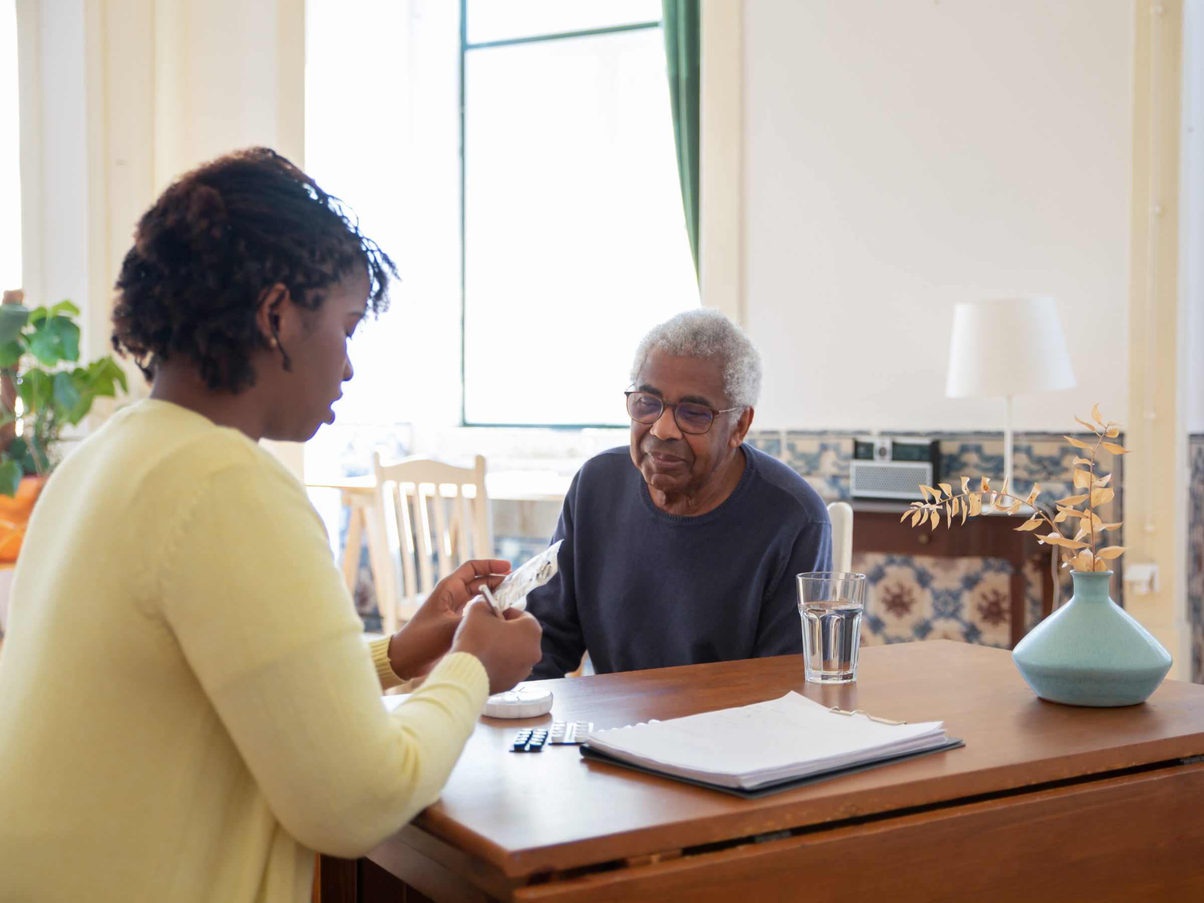 A caregiver sits with an older man at a table, reviewing his medication and offering support in a warm, home-like environment.