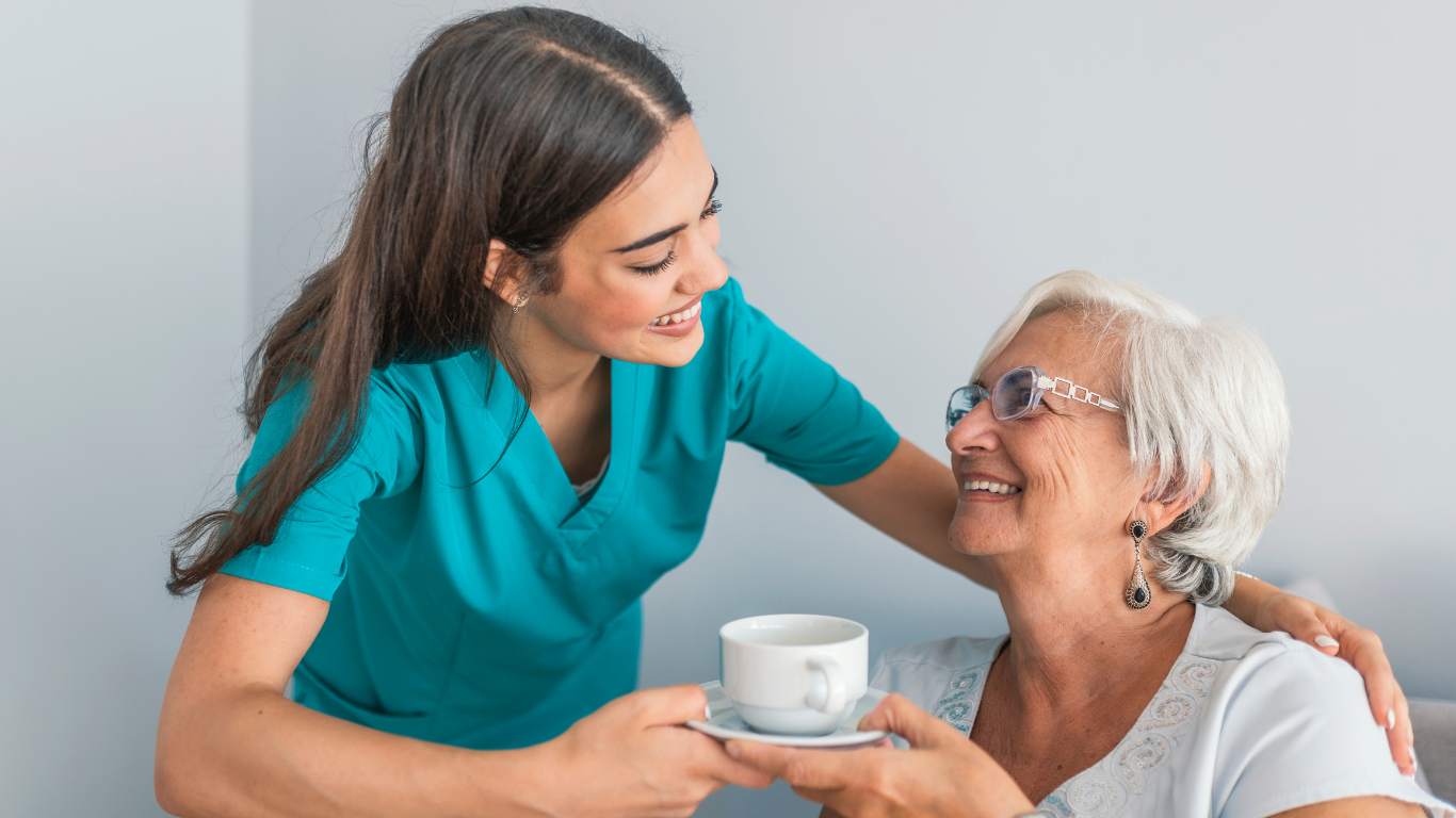 A caregiver shares a kind moment with an older woman, offering her a cup of tea and exchanging warm smiles.