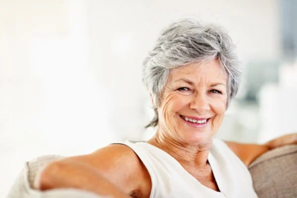A smiling older woman sits comfortably with her arms resting on a chair back, looking relaxed and content.