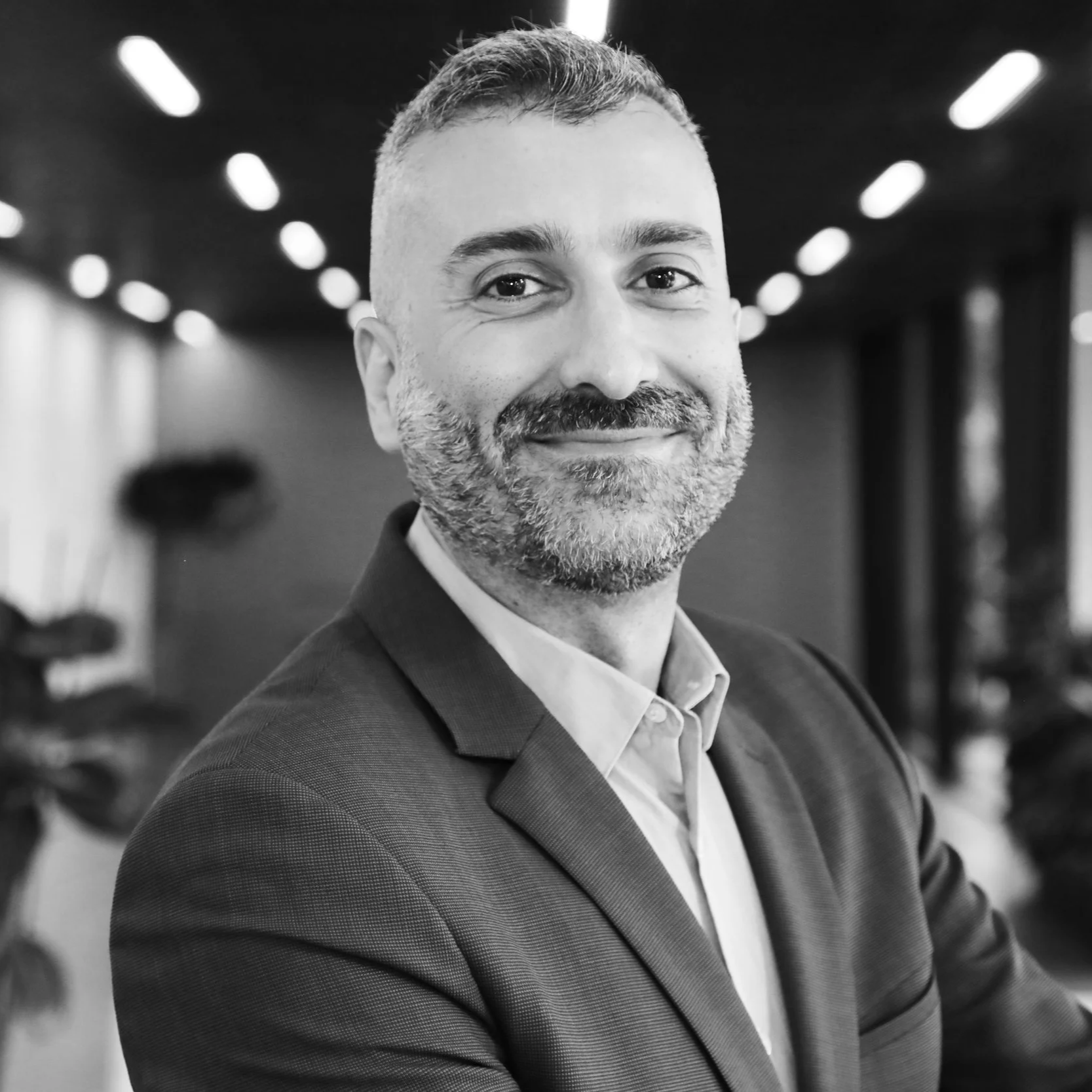 Black and white photo of a man in a suit smiling at the camera, with a blurred background of an indoor setting.