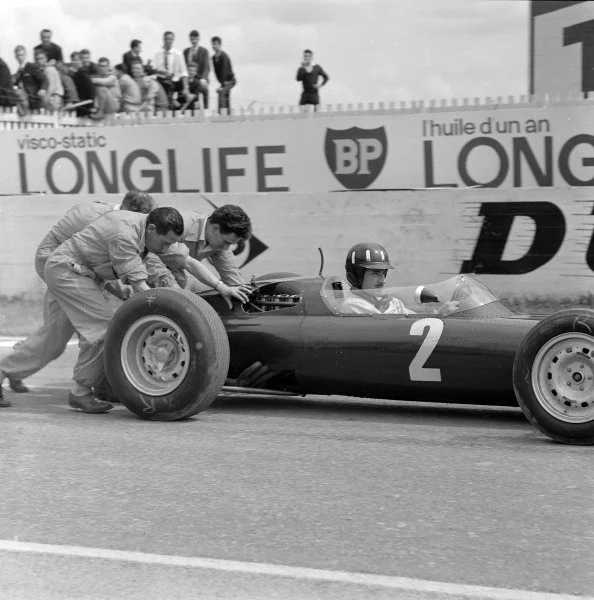 Two men pushing a race car with driver wearing helmet in pit stop, race track, and spectators in background.