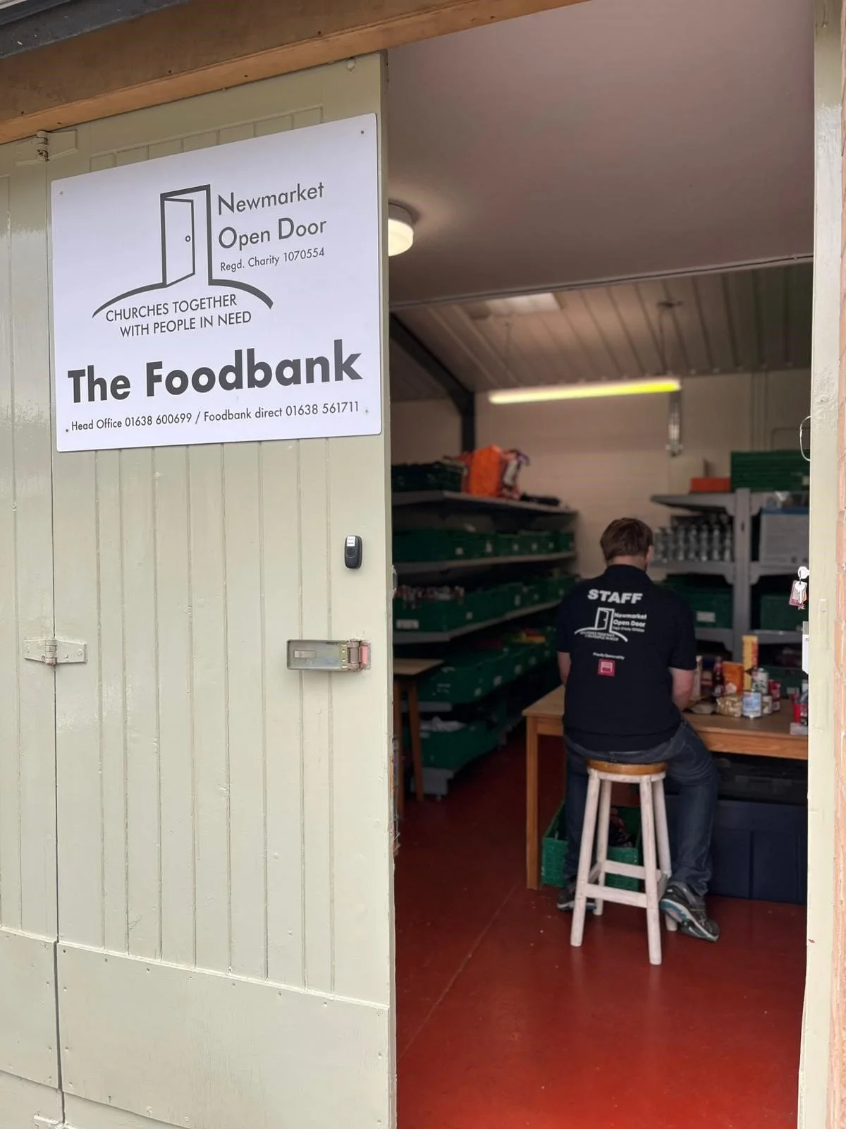 Open door at a food bank with a man sitting on a stool working at a table, wearing a black shirt with 'STAFF' on the back, inside a storage room with shelves of goods.