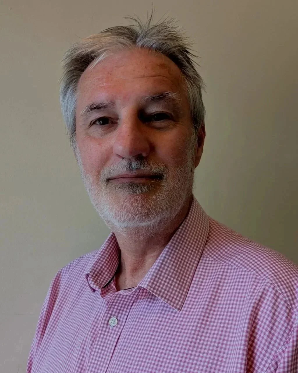 Portrait of an older man with gray hair and beard, wearing a pink and white checkered shirt, standing against a plain wall.