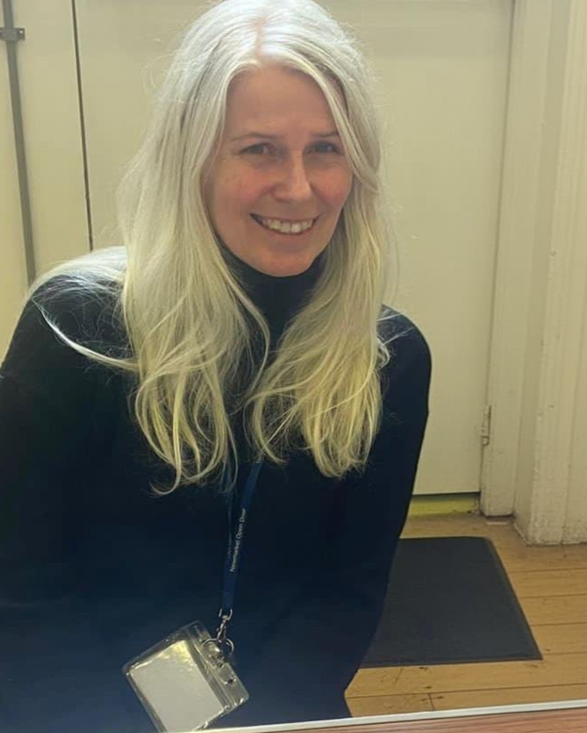 A woman with long platinum blonde hair, smiling, wearing a black jacket and a lanyard with an ID badge, sitting on a wooden floor near a white door.