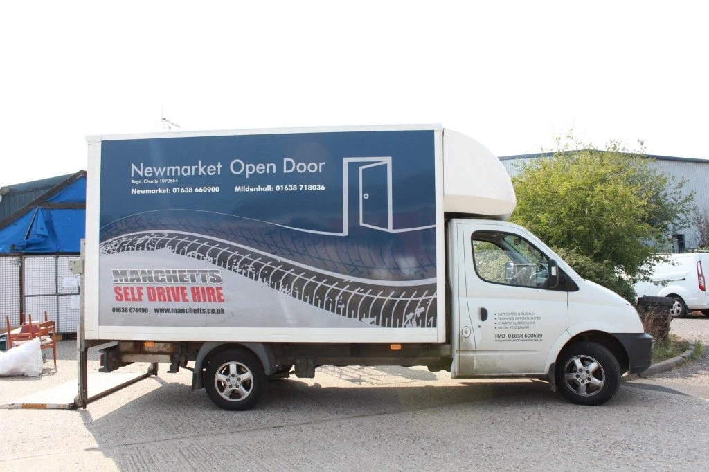 A white delivery truck with advertising for a self-drive hire tire company, parked outdoors near some trees and industrial buildings.