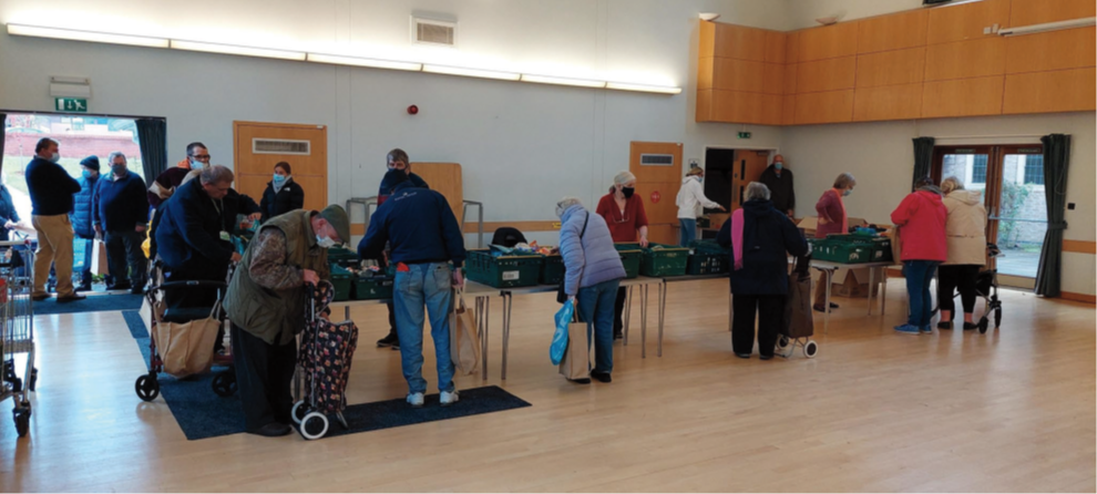 People shopping at a market stall inside a community center or hall, with some customers using shopping carts or trolleys.