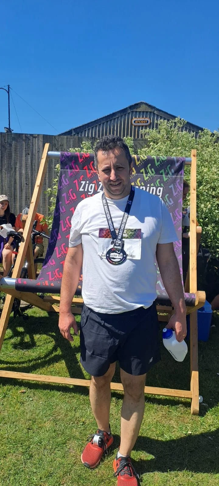 Man smiling with a medal around his neck after a race, holding a water bottle outdoors on a sunny day with other people in the background.