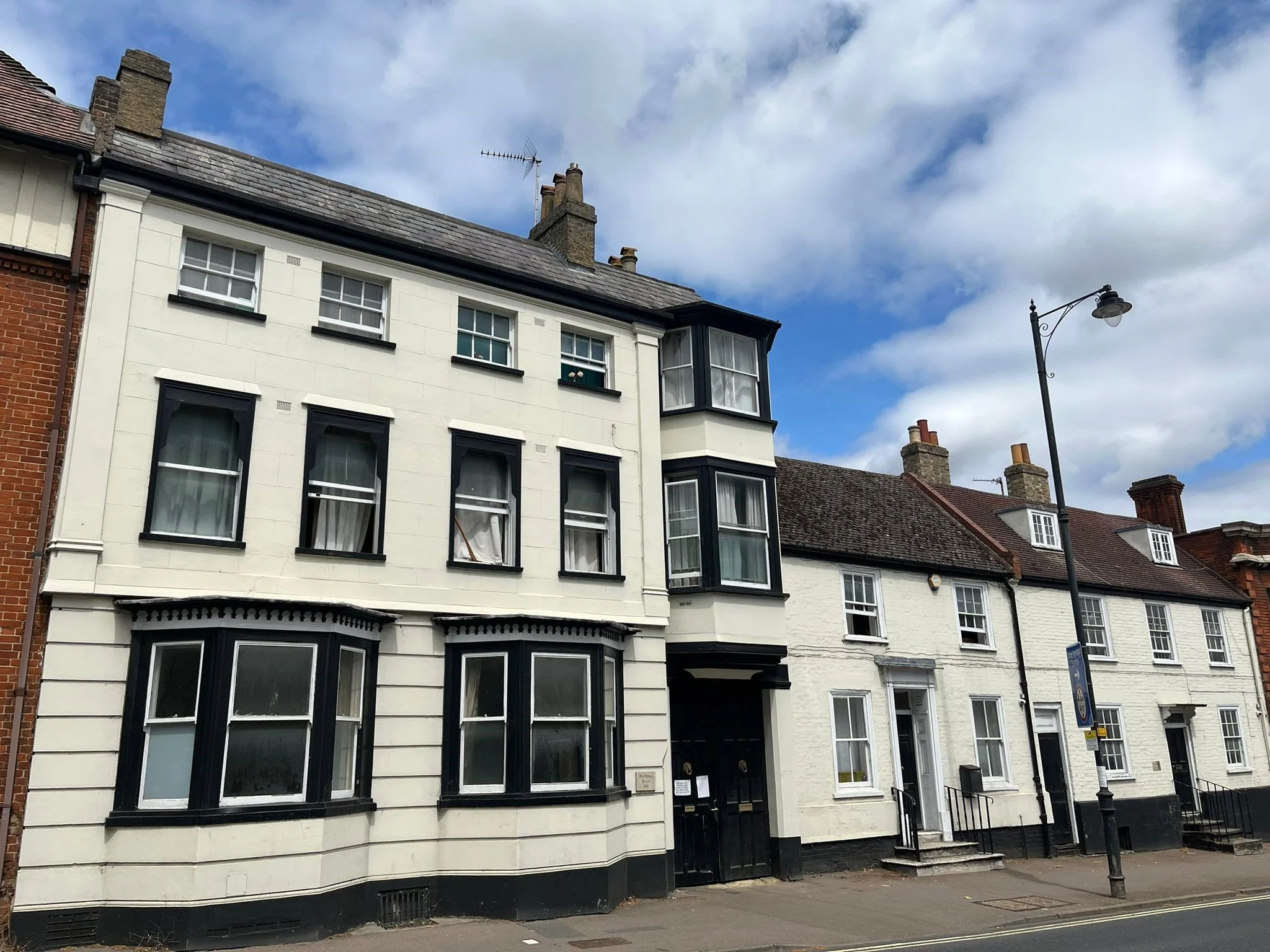 Row of traditional European-style houses with white and black facades, bay windows, and a streetlamp on the sidewalk under a partly cloudy sky.