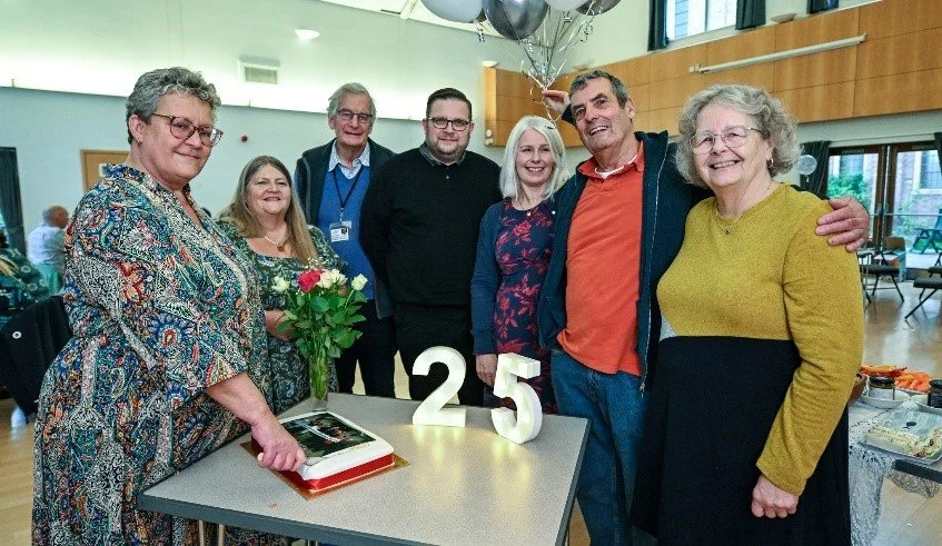 Group of seven adults celebrating in a room, with balloons, a bouquet of flowers, and illuminated numbers '25' on the table.