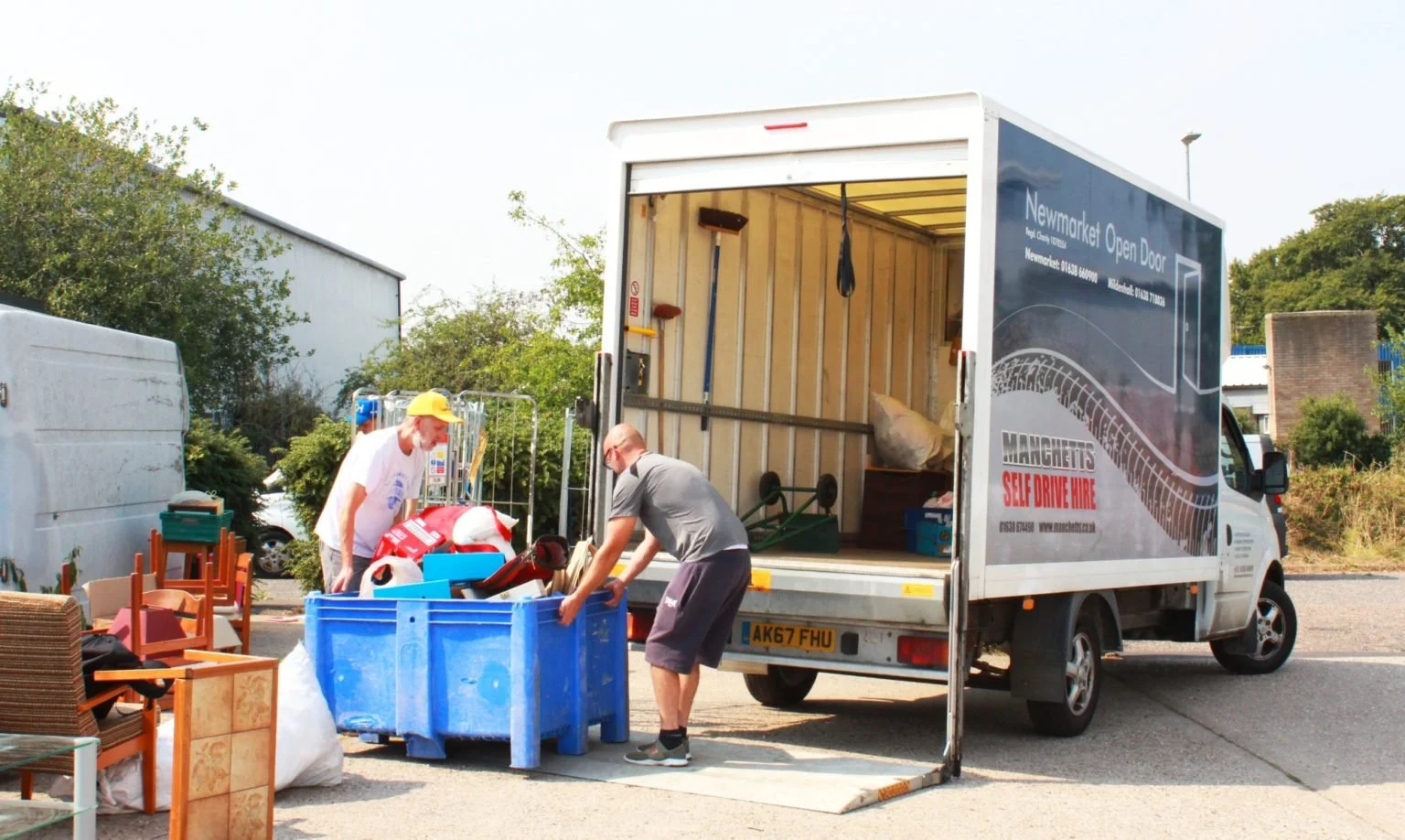 Two men unloading furniture and household items from a moving truck at a storage or moving site, with chairs and boxes nearby.