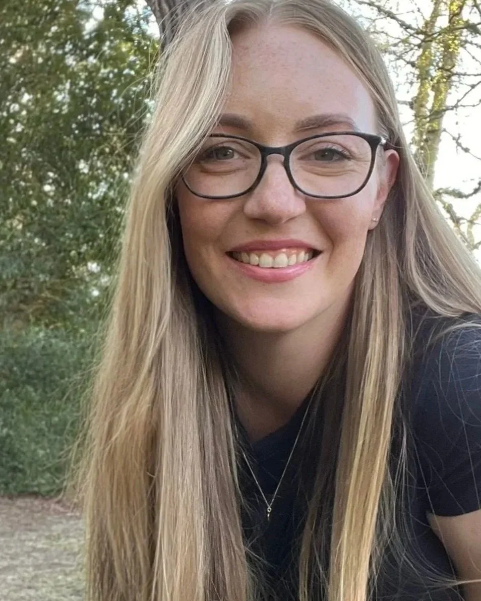A woman with long blonde hair wearing glasses, smiling outdoors with green trees in the background.