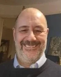 A smiling middle-aged man with a beard and mustache, wearing a collared shirt and sweater, indoors with a lamp and artwork in the background.