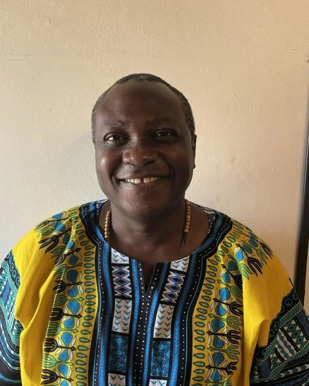 A smiling man wearing a colorful African dashiki with a beaded necklace, standing against a plain light-colored wall.