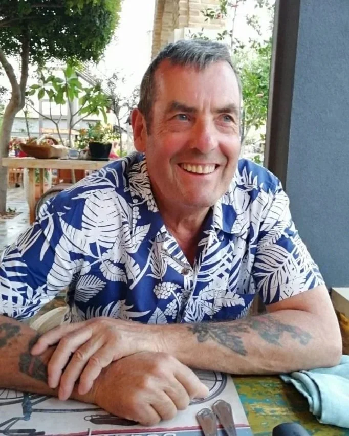A smiling man with tattoos on his arms is sitting at a table outdoors, wearing a blue and white tropical shirt, with plants and a patio in the background.
