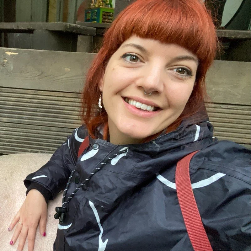 A woman with red hair, wearing a black jacket with white patterns, smiling outdoors with a wooden background.