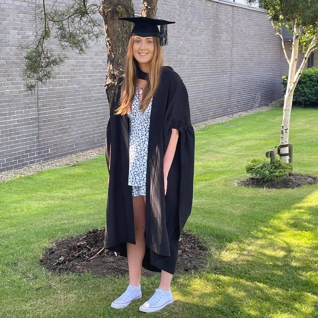 Young woman in graduation cap and gown standing on grass near a tree, smiling at camera.