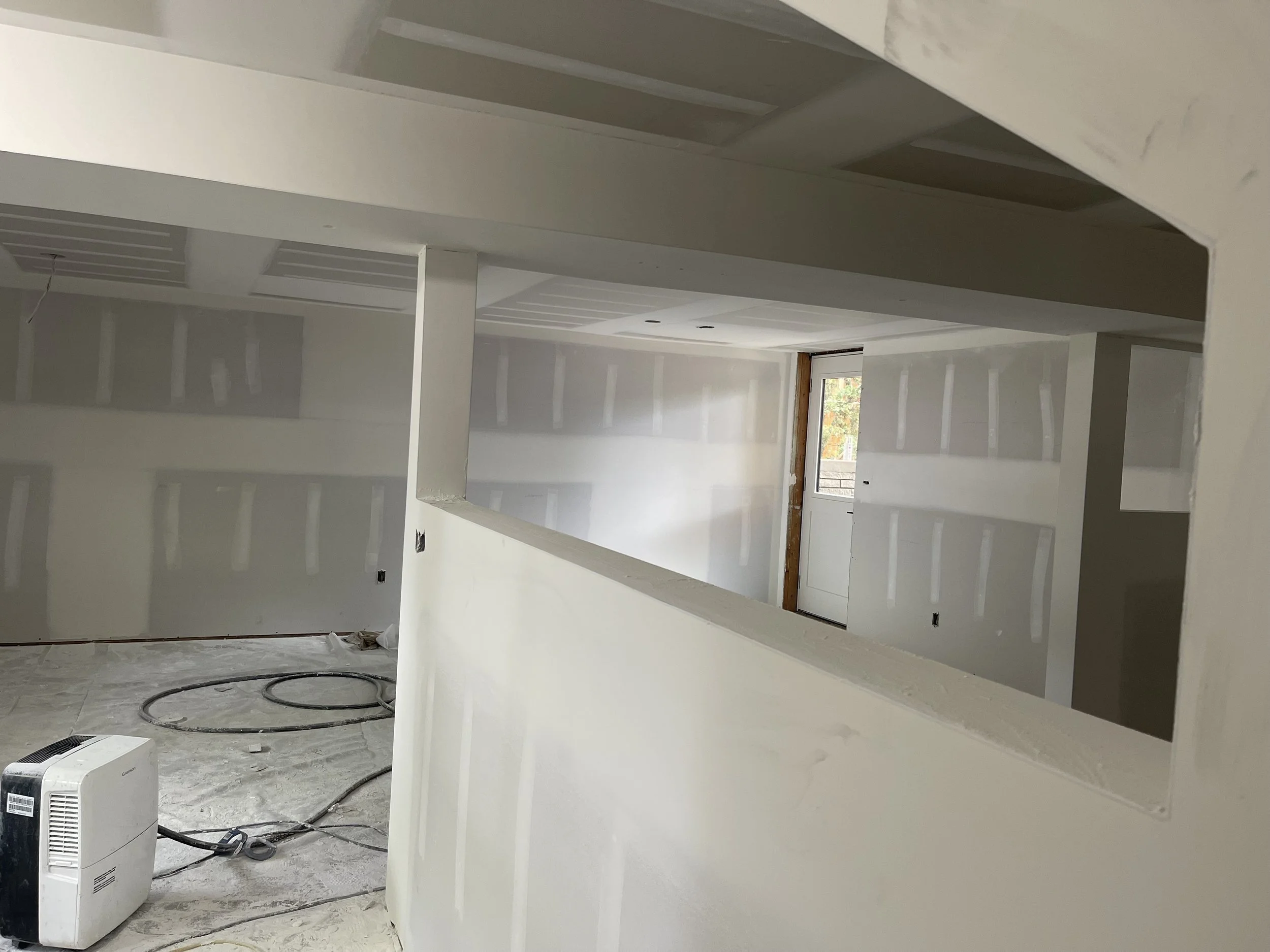 Interior of a building under construction or renovation, with white drywall on the walls and ceiling, an open window allowing natural light, and construction tools and materials on the floor.