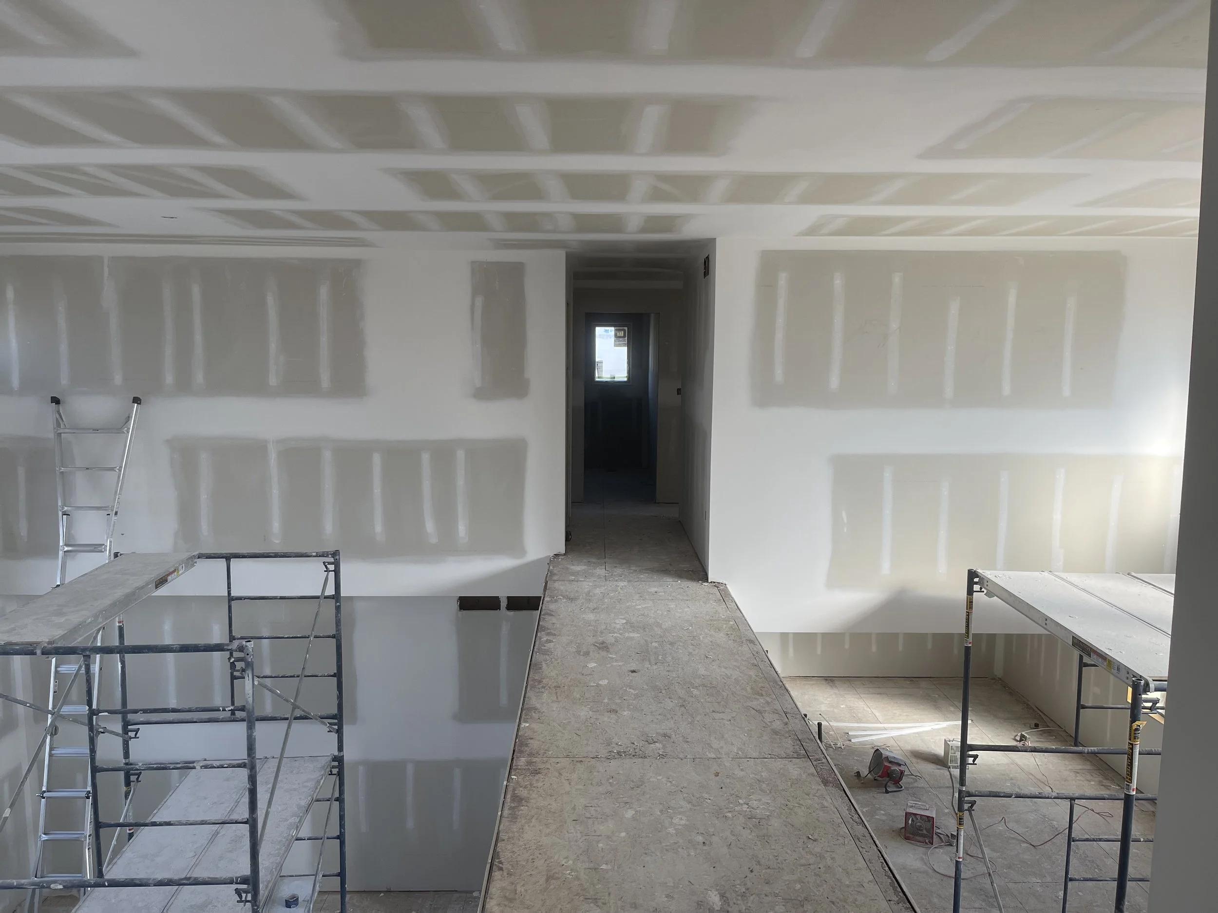 Interior of a house under construction with drywall on the walls and ceiling, scaffolding, construction tools, and an unfinished floor.