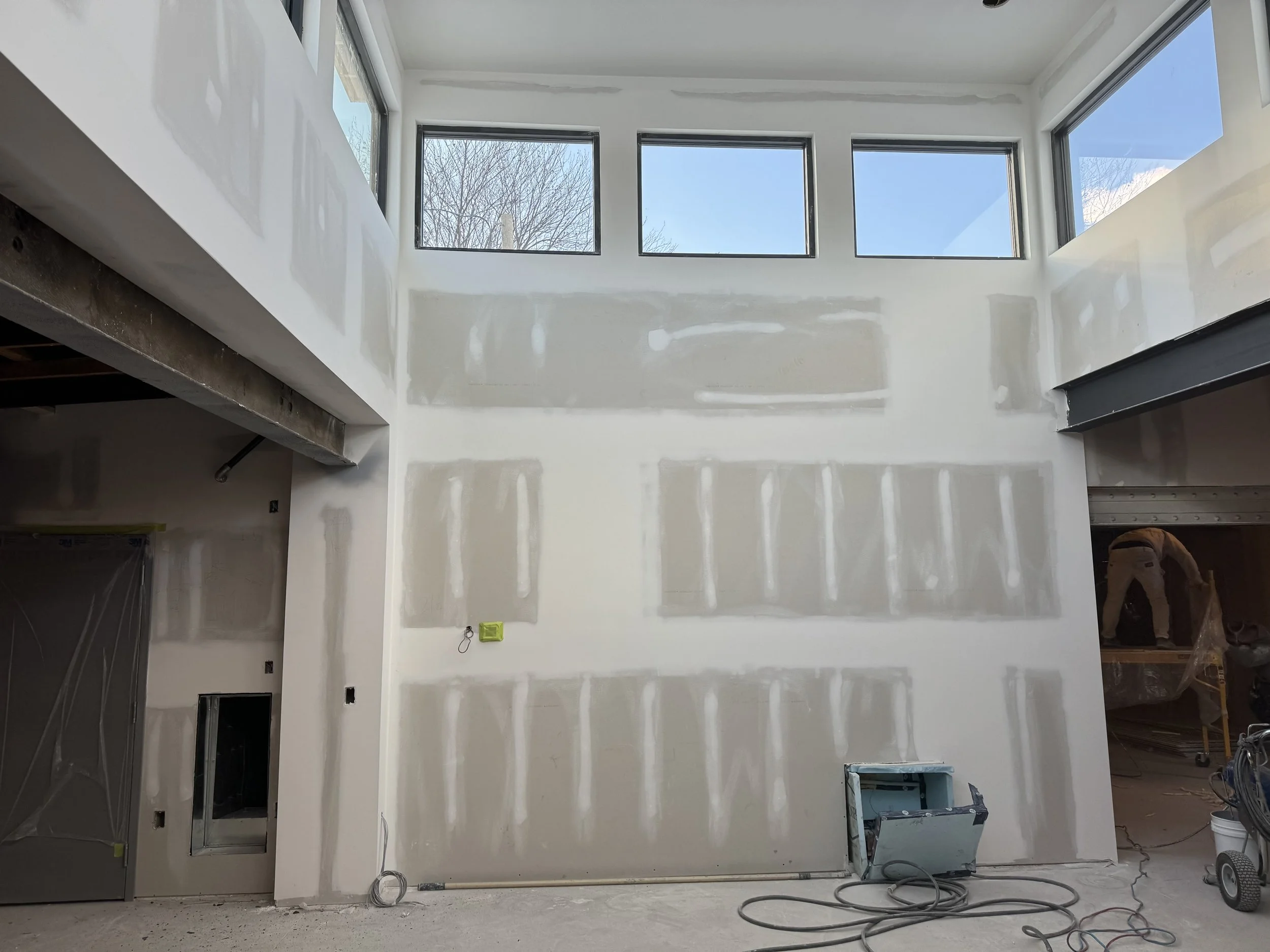 Interior of a house under construction or renovation, with unfinished drywall and hanging electrical wires, and large windows near the ceiling