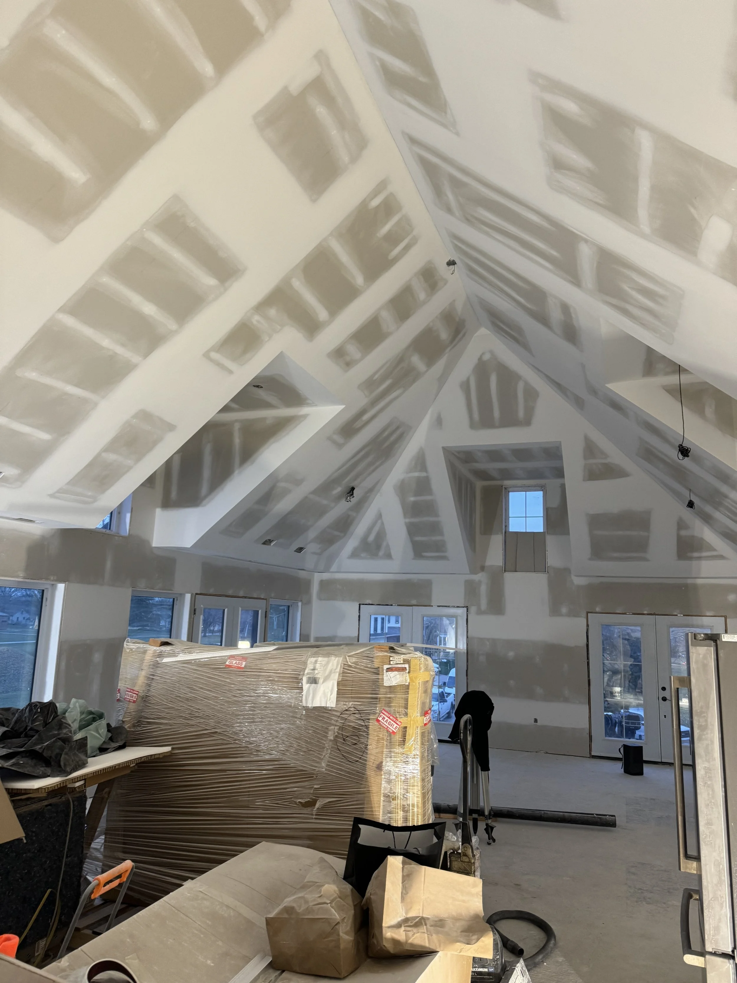 Room under construction with drywall installed and taped, ceiling with angled walls, large windows, and construction materials and tools scattered around.