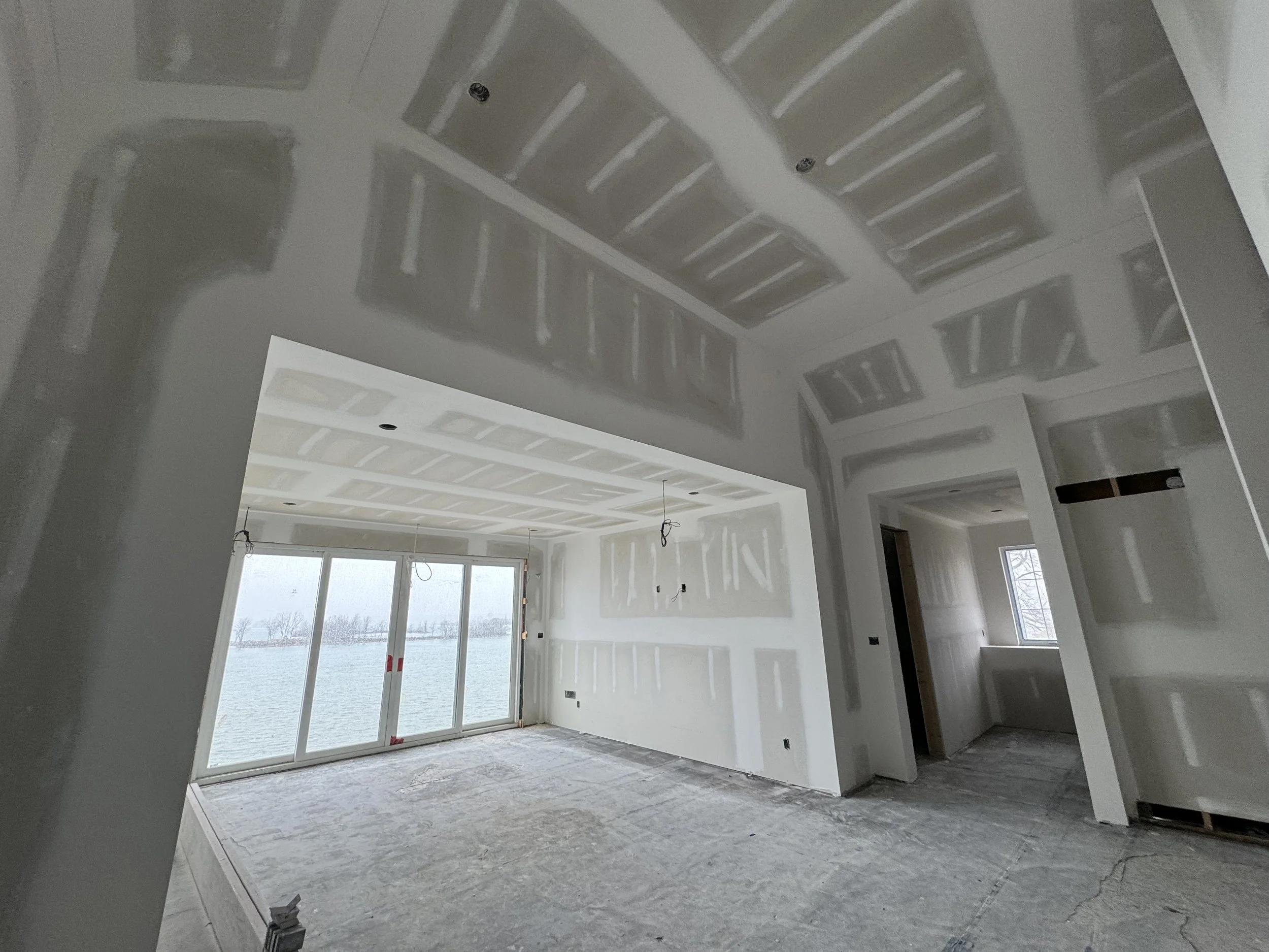 Interior of a house under construction with unfinished drywall, visible seams, and an open living space leading to a sliding glass door overlooking a body of water.