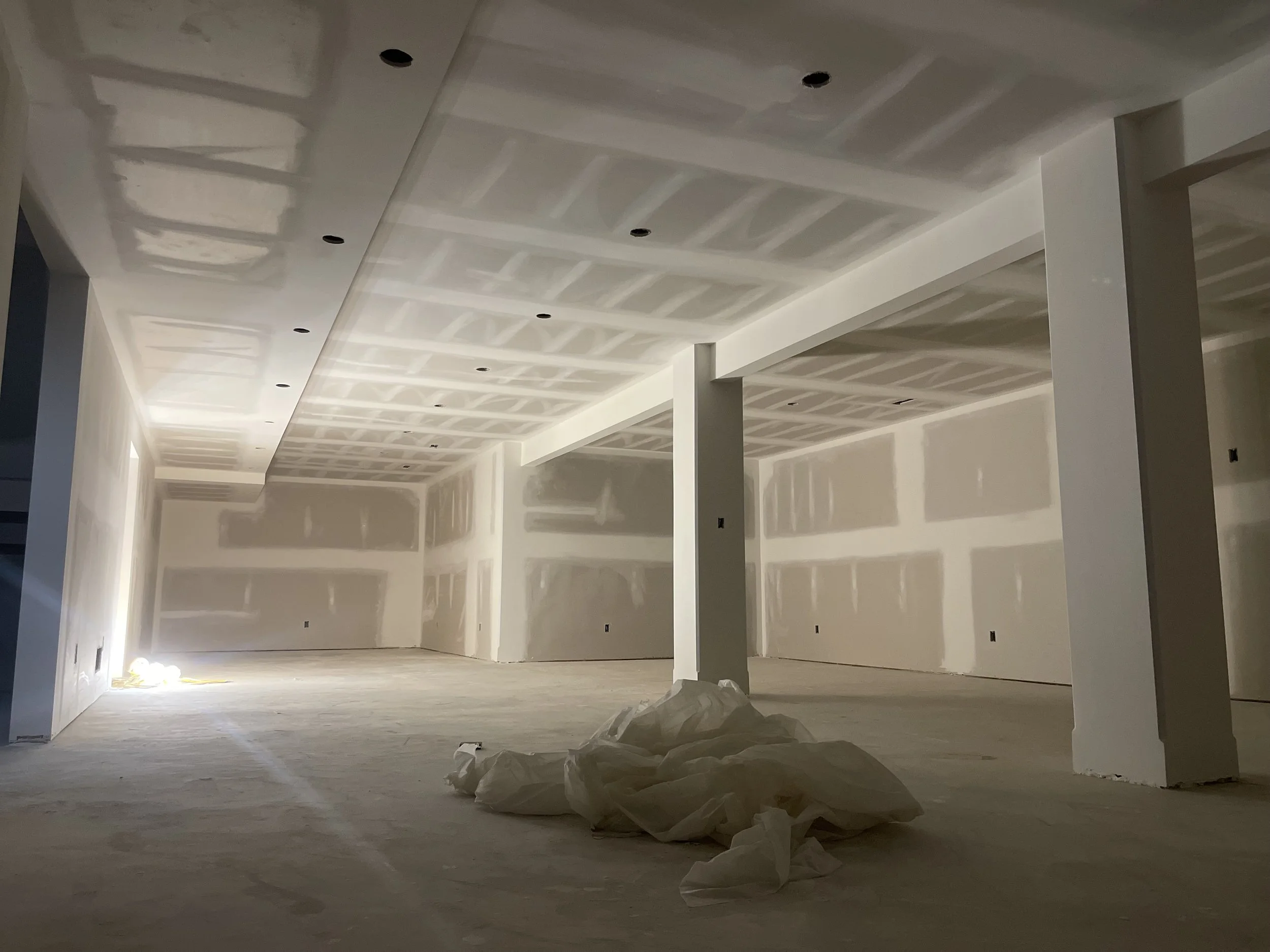 An unfinished room with drywall installed and ceiling texture in progress, with construction materials on the floor and no furniture yet.