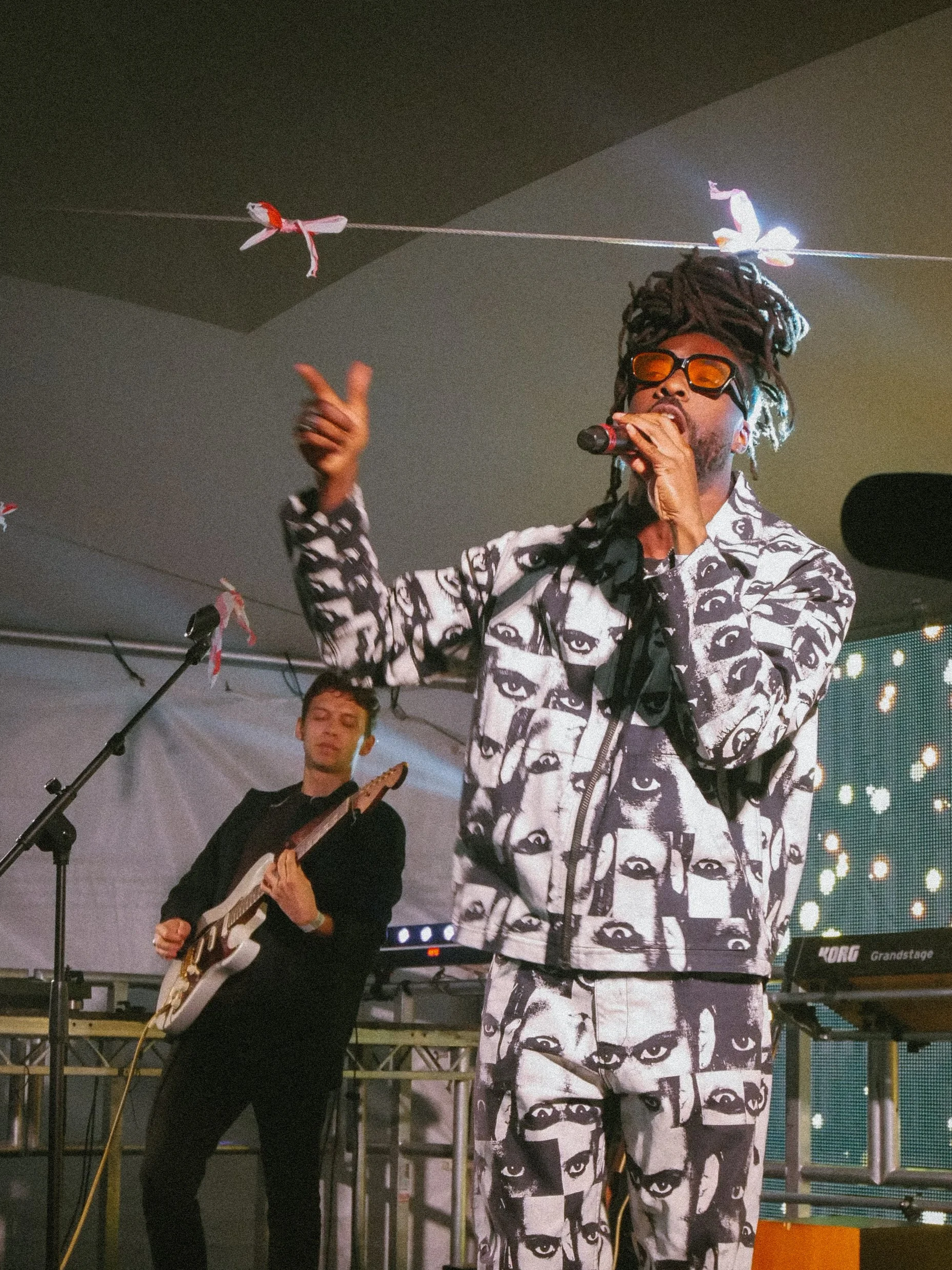 Singer with dreadlocks and sunglasses performing with a guitarist on stage, wearing a matching black and white outfit with eye patterns, in front of a LED screen and stage setup.