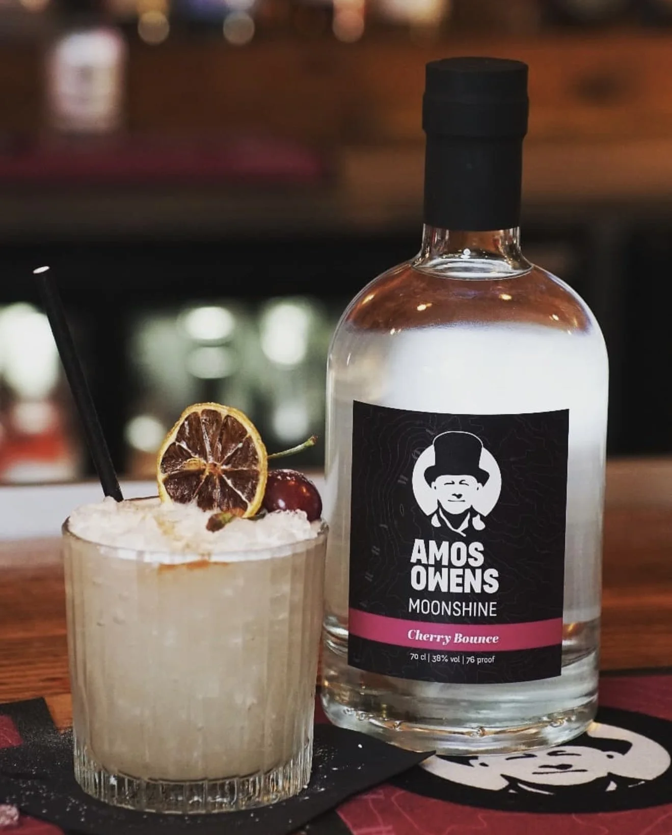 A cocktail garnished with a dried lemon slice, a cherry, and a black straw, placed next to a bottle of Amos Owens Moonshine Cherry Bounce on a bar counter.