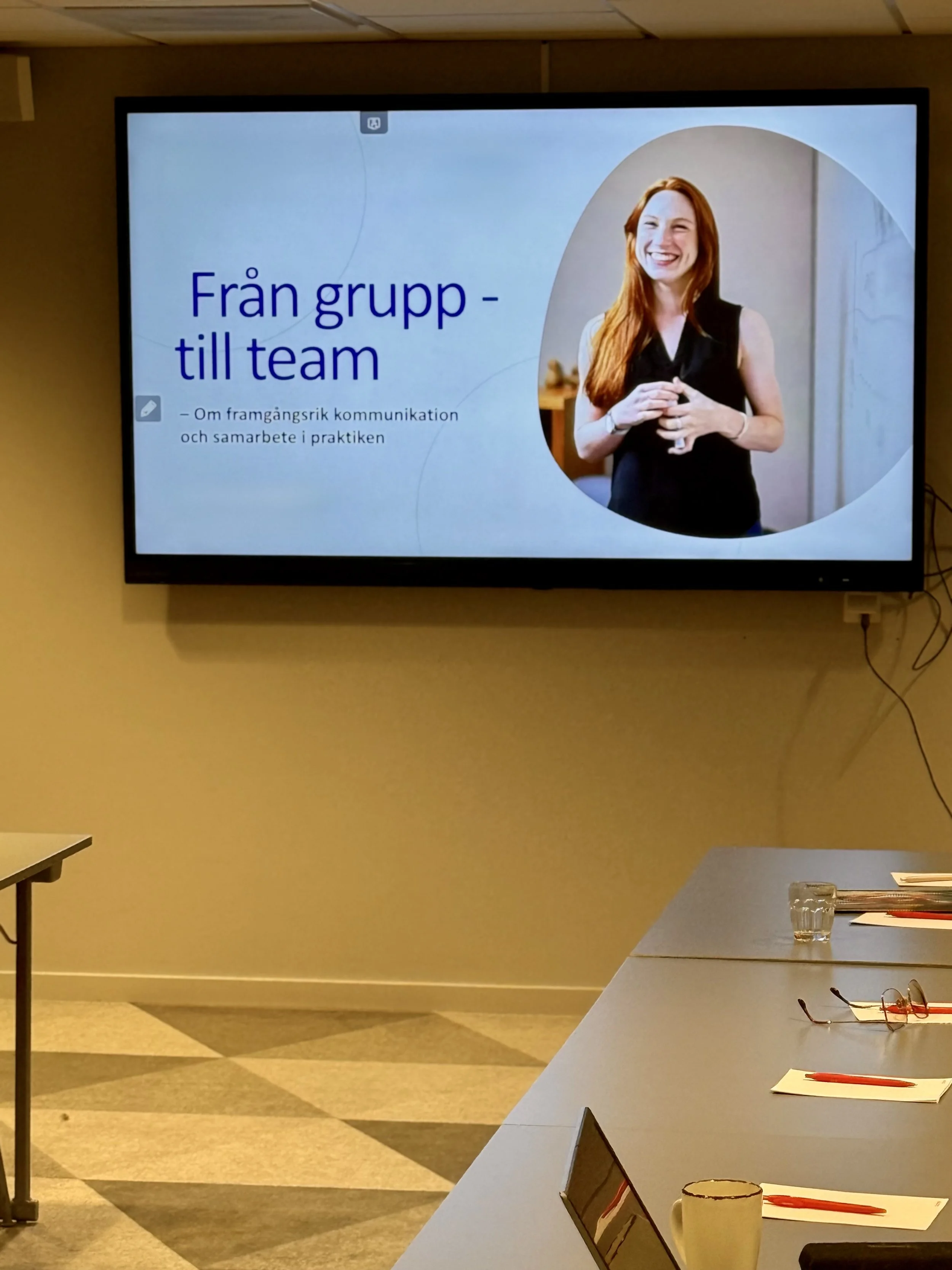 A presentation slide on a large screen shows a woman with long red hair, smiling and holding a glass. The slide title reads "Från grupp - till team" and a subtitle about successful communication and collaboration in practice.