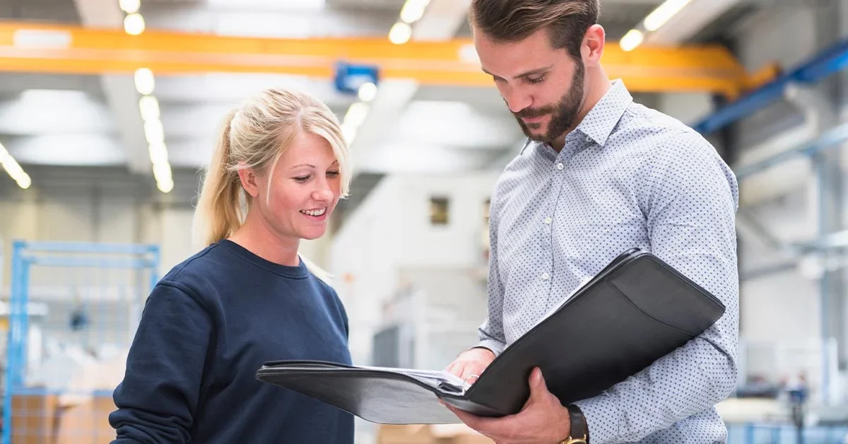 A man and a woman looking at a folder in an industrial or warehouse setting.