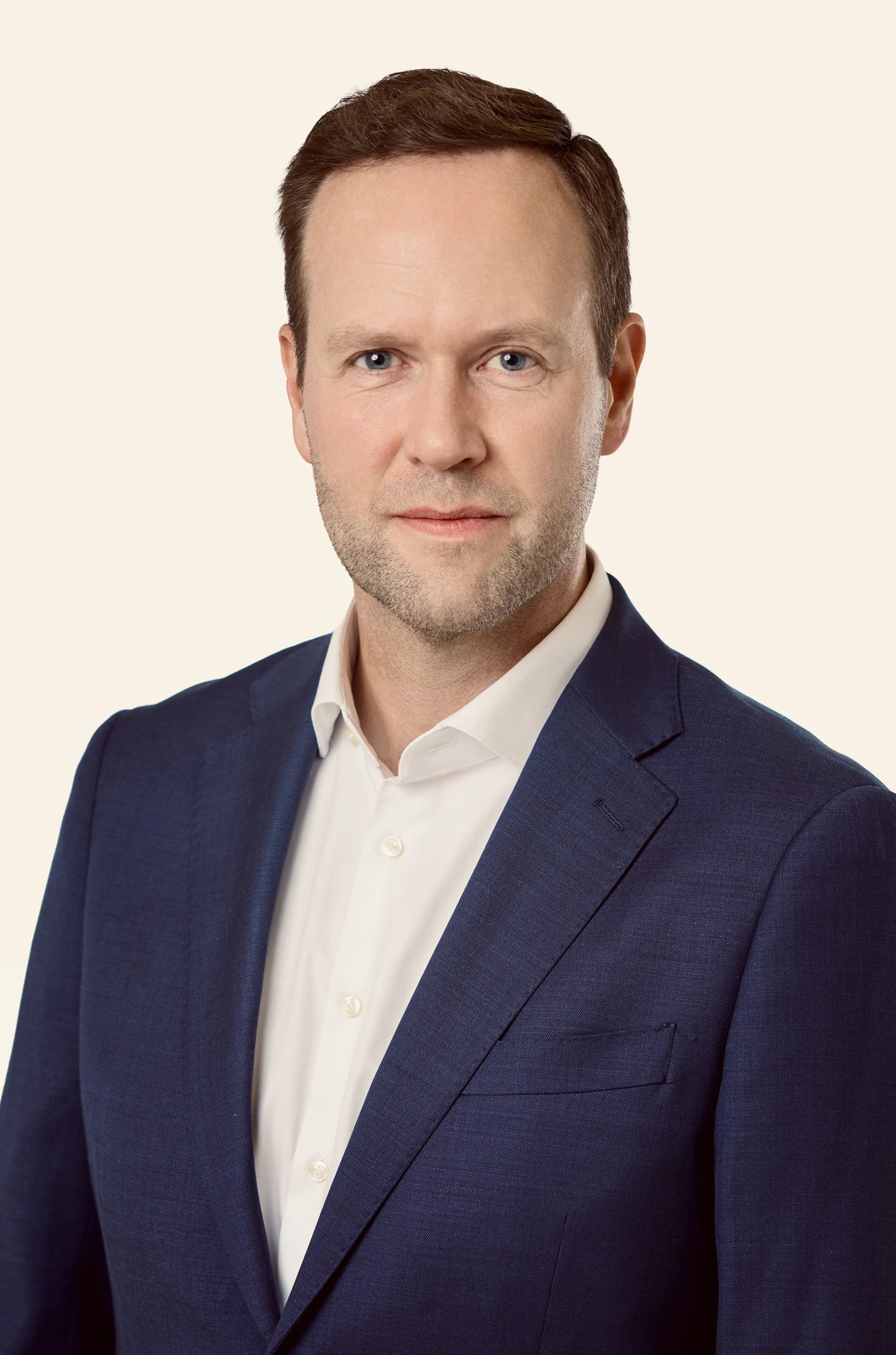 A professional man with light skin, short brown hair, wearing a navy blue suit and white dress shirt, looking directly at the camera against a plain light-colored background.