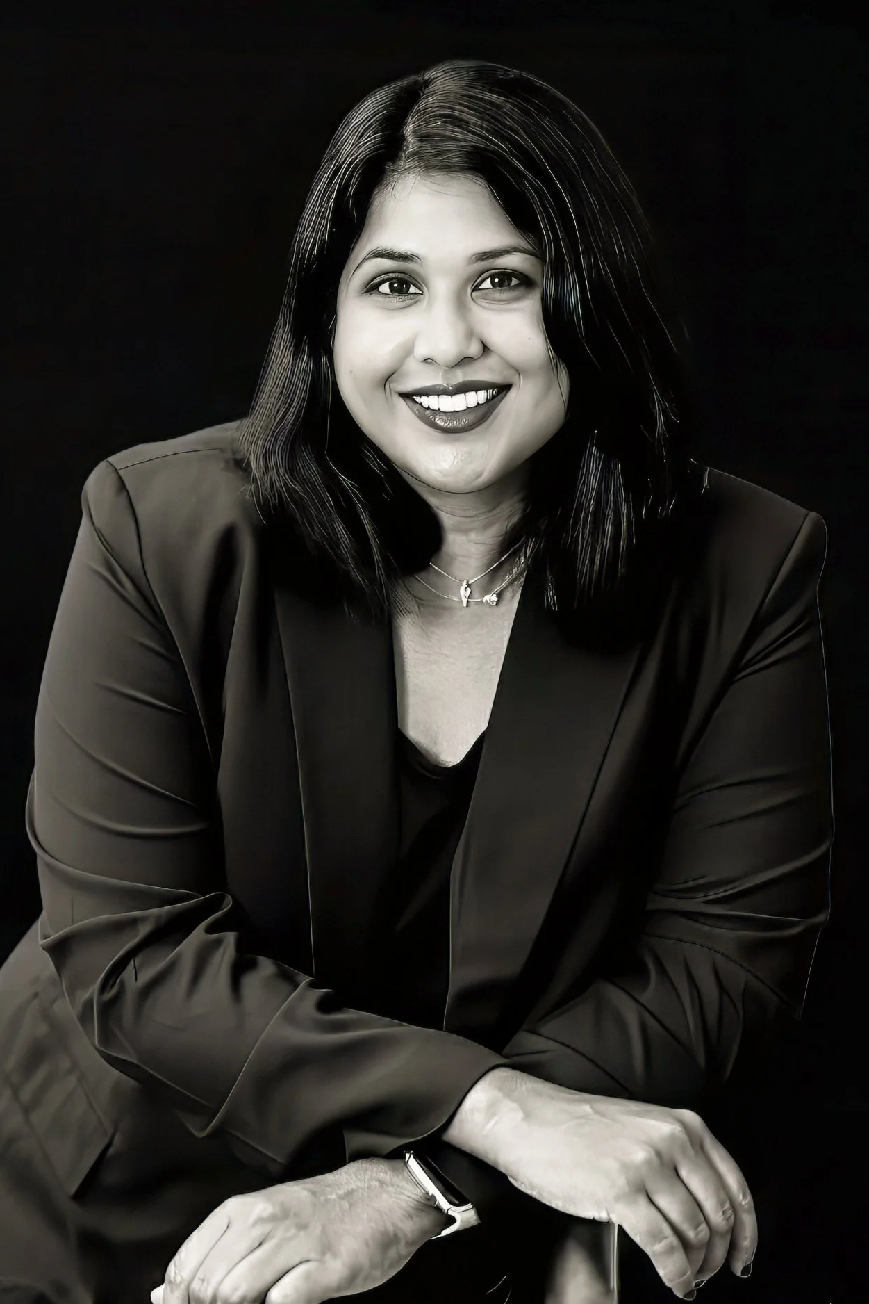 Black and white portrait of a smiling woman with shoulder-length dark hair, wearing a dark blazer, necklace, and watch, against a dark background.